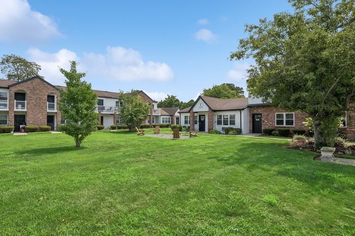 Charming apartment complex with brick facades and lush green lawns, offering a serene outdoor seating area under a clear blue sky.