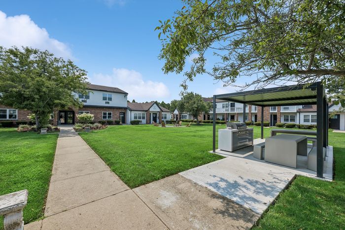 Charming apartment community with well-manicured lawns, outdoor seating, and a modern grilling area surrounded by scenic greenery.