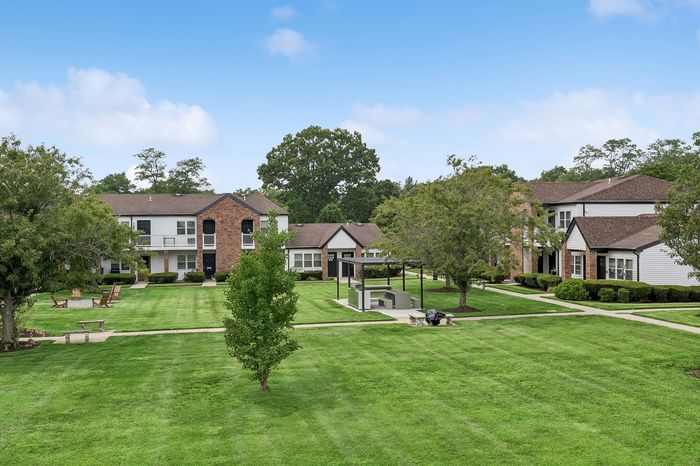 Charming apartment complex with lush green lawns, outdoor seating, and inviting walkways surrounded by mature trees under a bright blue sky.