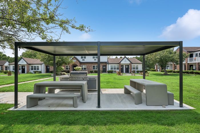Charming outdoor area with modern grill and picnic benches under a sleek pergola, surrounded by lush green lawns and stylish residential homes.