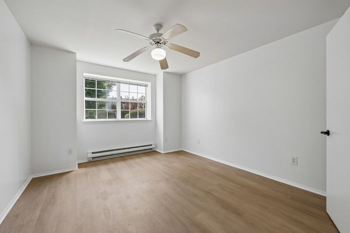 Bright, airy bedroom with sleek wood flooring, large window, and ceiling fan, perfect for a tranquil living space.