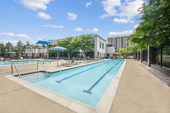Inviting outdoor pool area with sun loungers, bright blue umbrellas, and lush greenery in a modern apartment community.