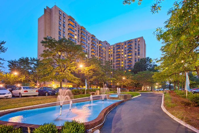Elegant high-rise apartment with illuminated windows, surrounded by lush greenery and a charming fountain, creating a serene urban retreat.