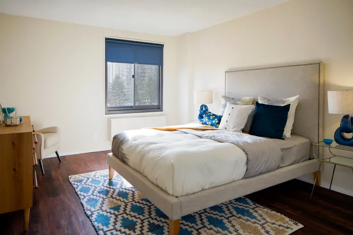 Bright and inviting bedroom with modern decor, featuring a plush bed, elegant side tables, and a soft patterned rug complementing the warm wooden flooring.