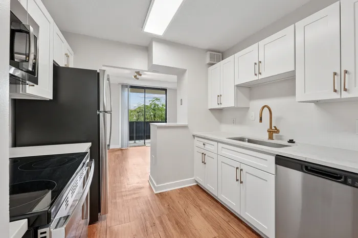 Modern kitchen with sleek white cabinets, stainless steel appliances, and elegant wood flooring leading to a scenic balcony view.