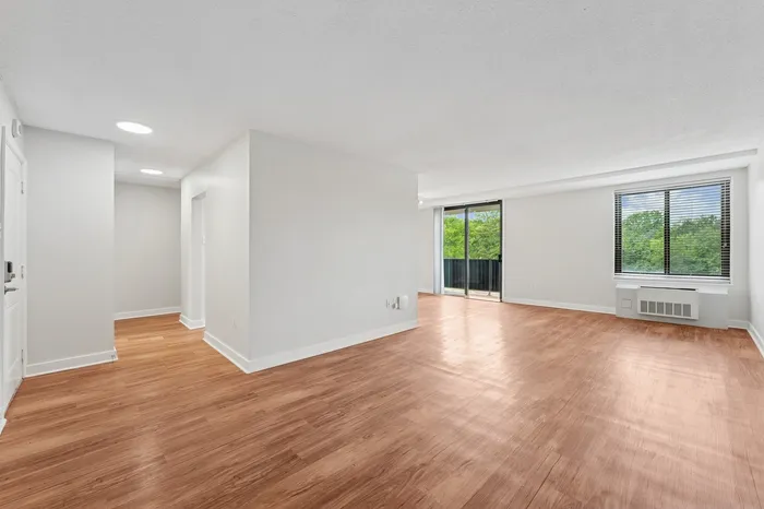 Bright and spacious living area with beautiful hardwood floors, large windows, and sliding doors leading to a balcony with lush green views.