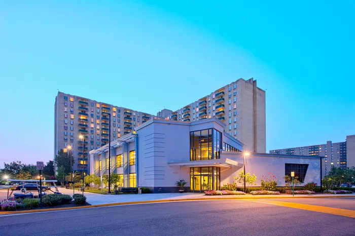 Modern high-rise apartment building with expansive glass-fronted entry, beautifully lit at dusk, surrounded by lush landscaping and ample parking.