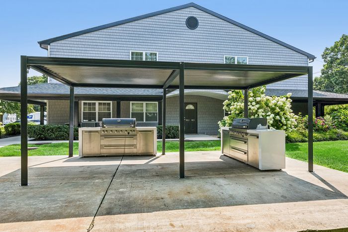 Modern outdoor BBQ area with sleek grills under a stylish pergola, set against a charming residential backdrop surrounded by lush greenery.