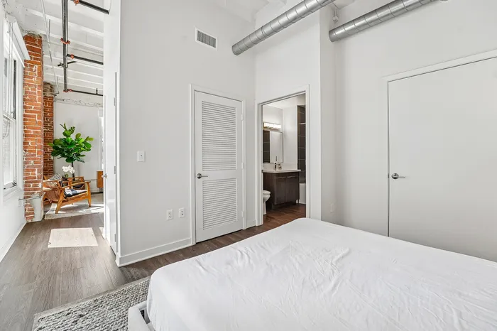 Modern bedroom with white walls and exposed brick, featuring a sleek en-suite bathroom, and contemporary decor in an open living area.