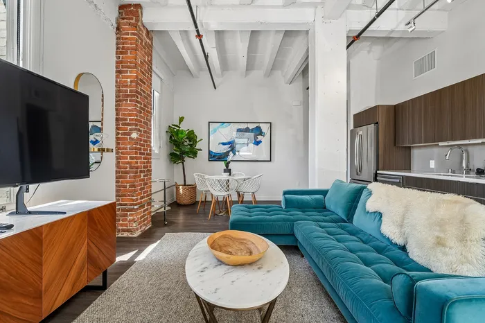 Modern open-concept living space featuring an elegant teal sofa, sleek kitchen with stainless steel appliances, and exposed brick accents for a stylish urban vibe.