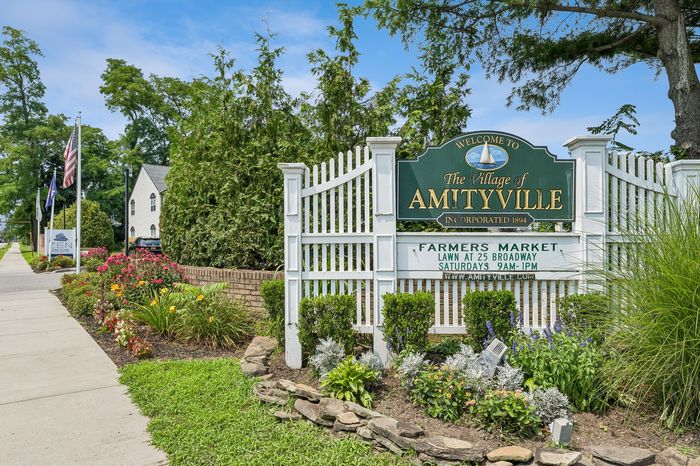 Charming entrance to The Village of Amityville with a welcoming sign, vibrant flowers, and lush greenery.