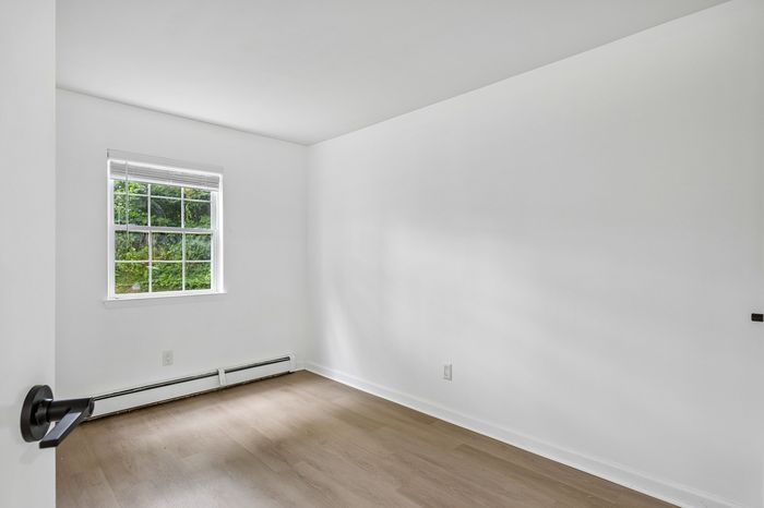 Bright and airy room with fresh white walls, wood flooring, and a large window offering serene outdoor views.