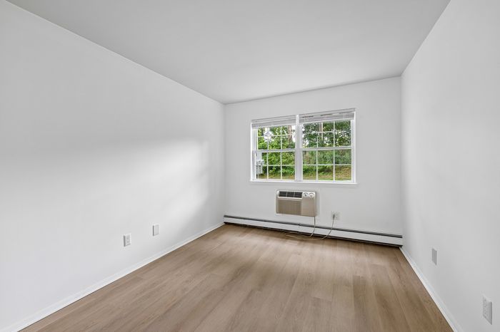 Bright and airy room with modern hardwood floors, large window with garden view, and an efficient wall-mounted air conditioner.