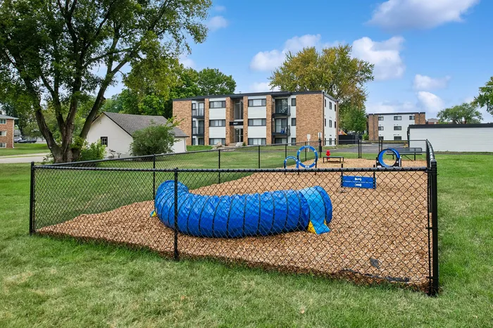Inviting apartment complex highlighting a vibrant dog park with agility equipment, set within lush greenery and modern buildings.