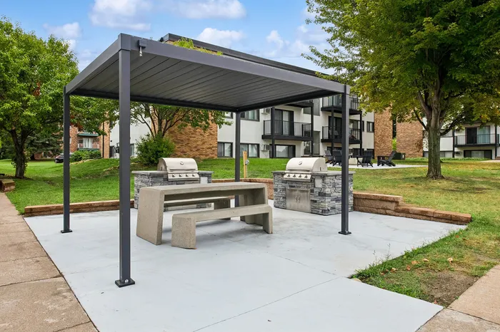 Inviting outdoor BBQ area with modern grills under a stylish pergola, surrounded by lush greenery and apartment buildings.