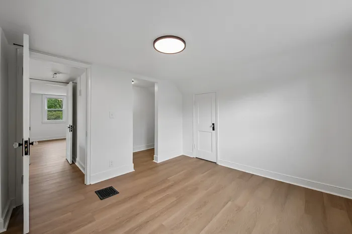 Elegant room with pristine white walls, modern ceiling light, and warm wooden flooring, offering abundant natural light through a nearby window.