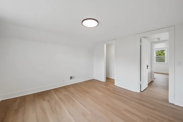 Bright and airy room with elegant wooden flooring and a modern ceiling light fixture, leading to a spacious area with a large window.