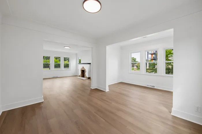 Bright and airy open living space with elegant hardwood floors and a charming fireplace, highlighting large windows with lush garden views.