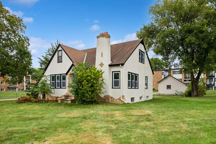 Charming white cottage with a classic gable roof set amidst lush green lawns, featuring a quaint front porch and surrounded by mature trees under a clear blue sky.
