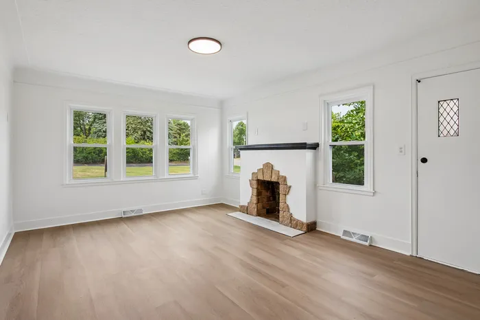 Bright and airy living room with large windows offering lush green views and a charming stone fireplace.