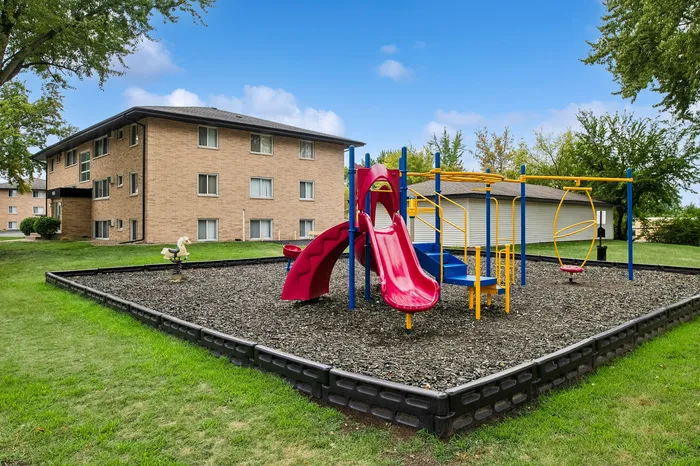 Inviting apartment complex with vibrant children's playground area, featuring a bright red slide and colorful climbing structures, surrounded by lush greenery.