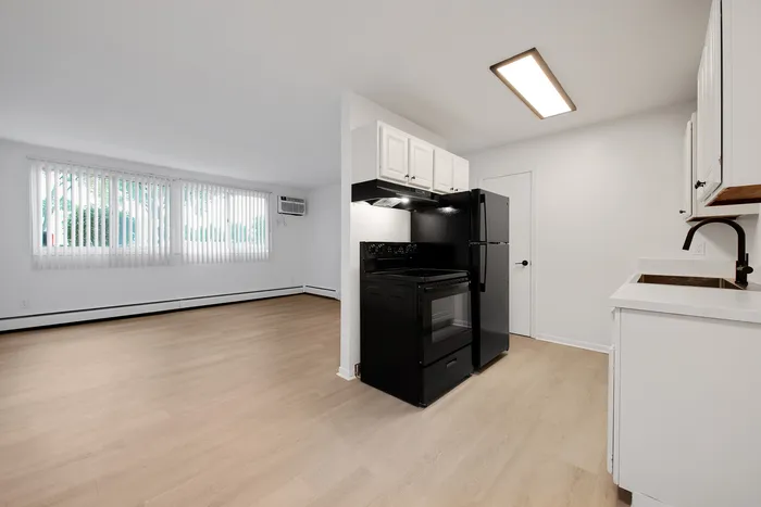 Bright and spacious apartment interior featuring an open-plan layout with light wood flooring, a sleek black kitchen with modern appliances, and large windows with vertical blinds.