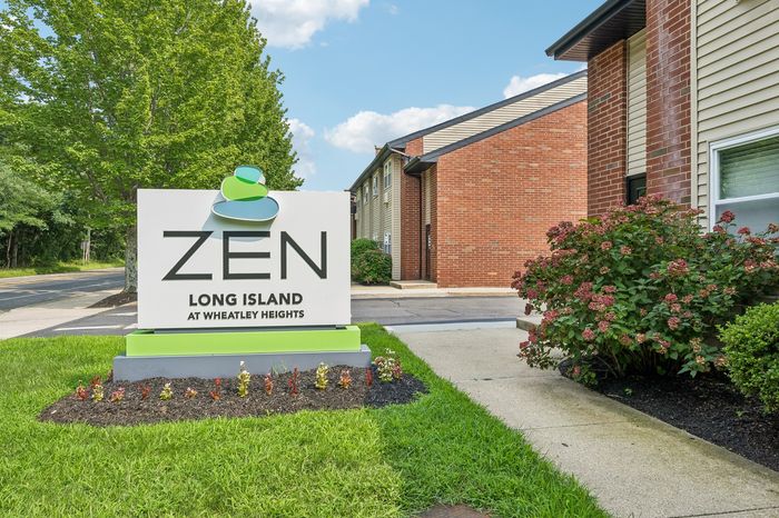 Welcoming entrance to Zen Long Island at Wheatley Heights, featuring vibrant greenery and a modern building facade under a clear blue sky.