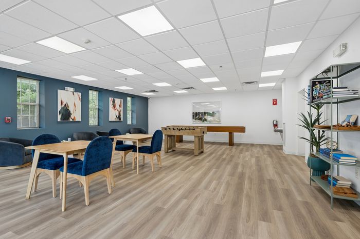Modern community recreation room featuring elegant wooden flooring, cozy seating, stylish blue chairs, decorative artwork, and engaging game tables.