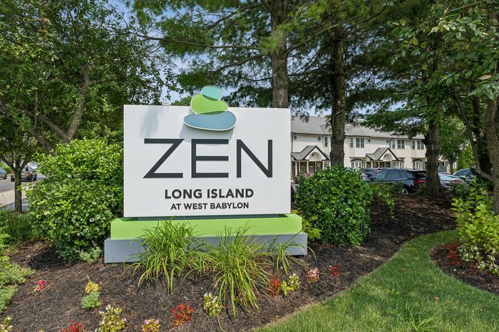 Welcoming entrance sign to Zen Long Island at West Babylon, surrounded by lush greenery and charming residential buildings.