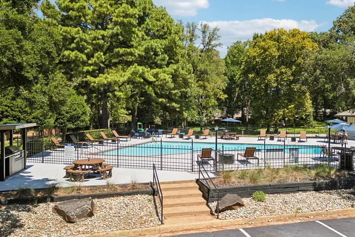 Tranquil outdoor pool area surrounded by lush trees with sun loungers and shaded picnic spots, offering a serene escape.