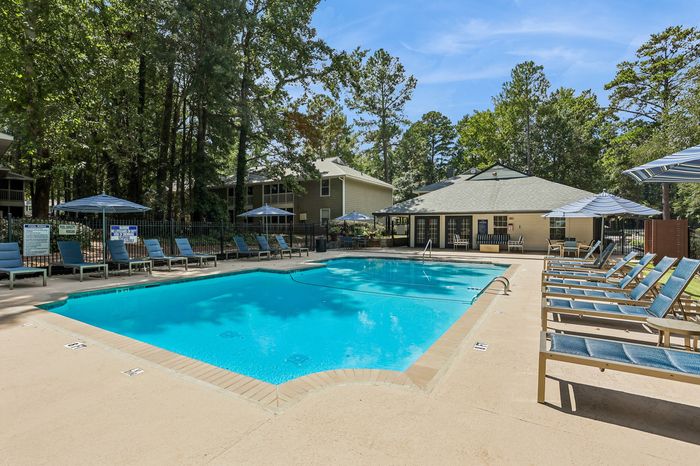 Inviting outdoor pool area with luxurious lounge chairs, sunshaded tables, surrounded by lush greenery and modern apartment buildings.