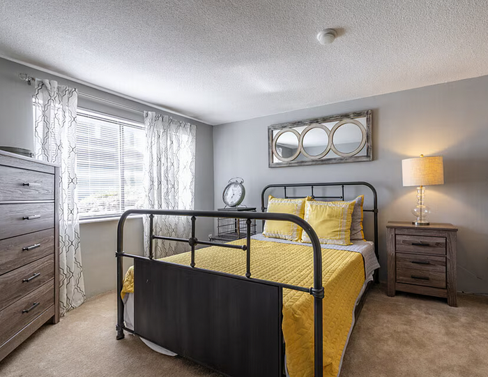 Bright and cozy bedroom featuring a modern metal bed with vibrant yellow bedding, elegant wooden furniture, and ample natural light through large windows.