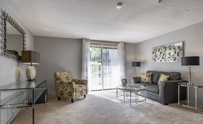 Bright and modern living room with elegant furniture, featuring a plush gray sofa, patterned armchair, and sleek glass tables, complemented by stylish decor and large windows with sheer curtains.