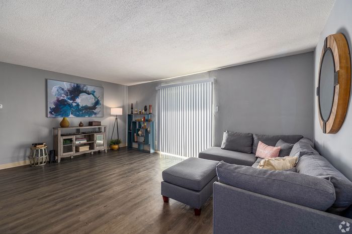 Spacious living room with sleek hardwood floors, large grey sectional sofa, modern art piece, and ample natural light through vertical blinds.
