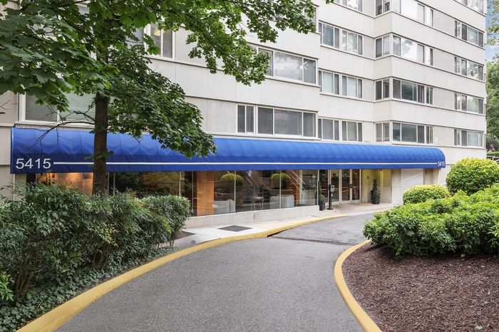 Charming apartment building entrance with a blue awning, lush greenery, and inviting pathway, perfect for urban living.