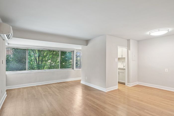 Bright and airy living space with elegant hardwood floors, large window with lush greenery view, and sleek kitchen entrance.