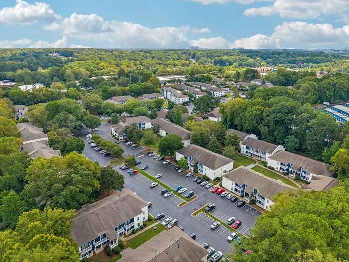 Aerial view of charming apartment complex nestled in lush greenery, showcasing well-maintained buildings, ample parking, and serene surroundings.