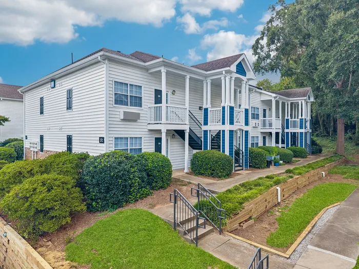 Charming two-story apartment building with elegant white siding, inviting balconies, and lush landscaping, under a bright blue sky.