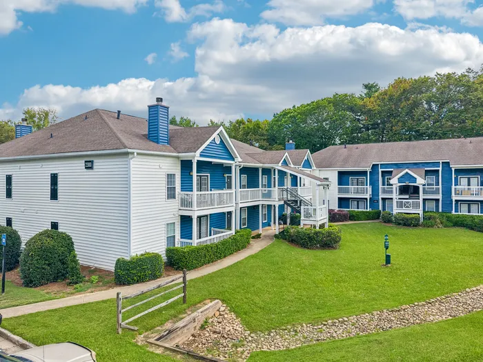 Charming blue and white apartment complex with lush green lawns and inviting balconies, set against a backdrop of vibrant trees and a bright sky.