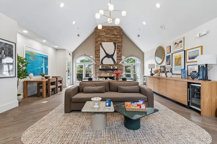 Inviting living space with high vaulted ceilings, a modern chandelier, and a cozy stone fireplace, flanked by arched windows offering abundant natural light. Stylish decor and a spacious sofa create a perfect blend of comfort and elegance.