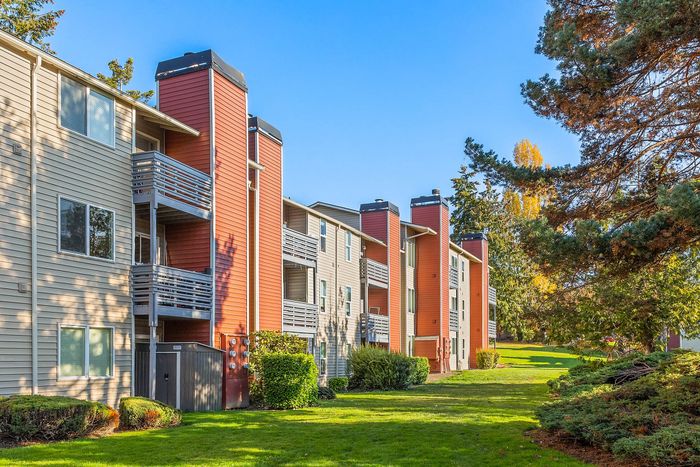 Charming apartment complex with red and beige exteriors, featuring spacious balconies surrounded by lush greenery and blue skies.