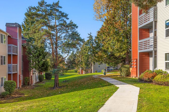 Charming apartment complex with vibrant red and cream exteriors, surrounded by lush greenery and towering trees, featuring a scenic sidewalk perfect for leisurely walks.
