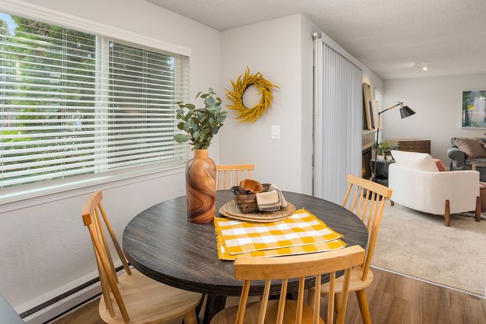 Cozy dining area in a bright open-plan apartment featuring a modern round table with elegant wooden chairs, large window with lush greenery view, and a contemporary living room accentuated by chic decor and ample natural light.