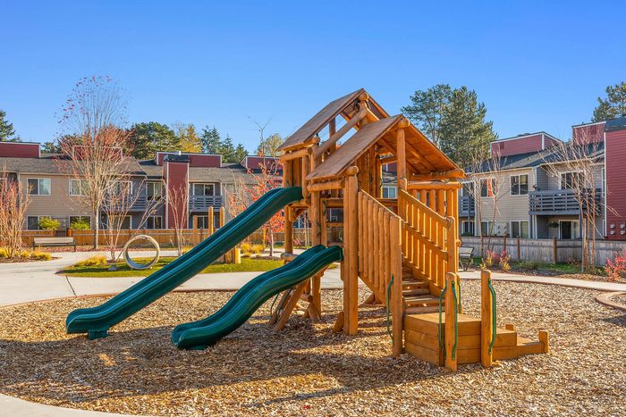 Charming outdoor playground featuring a wooden play structure with slides, surrounded by vibrant autumn foliage, in a serene apartment community setting.
