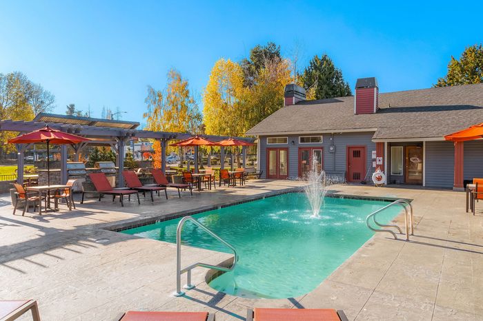 Inviting outdoor pool area with sun loungers, tables with umbrellas, and a charming clubhouse, surrounded by lush autumn trees.