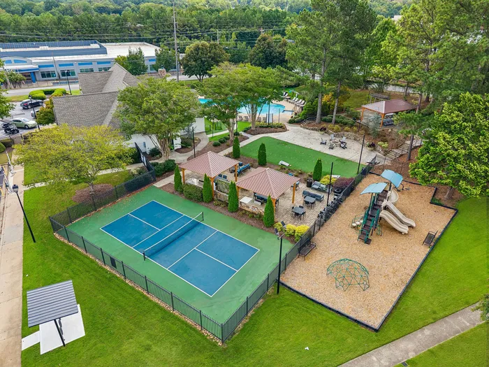 Captivating aerial view of a community recreation area featuring a tennis court, playground with slides, picnic gazebos, and a pool surrounded by lush greenery.