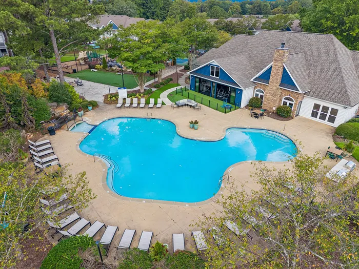 Inviting aerial view of a sparkling, resort-style swimming pool surrounded by lush greenery and comfortable lounge chairs, adjacent to a charming clubhouse.