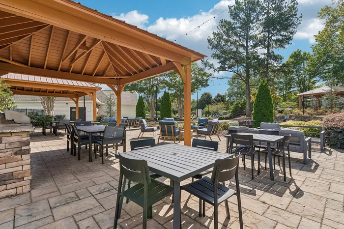 Charming outdoor patio with elegant seating under a wooden pergola, surrounded by lush greenery and sunshine; perfect for a relaxing gathering or alfresco dining.