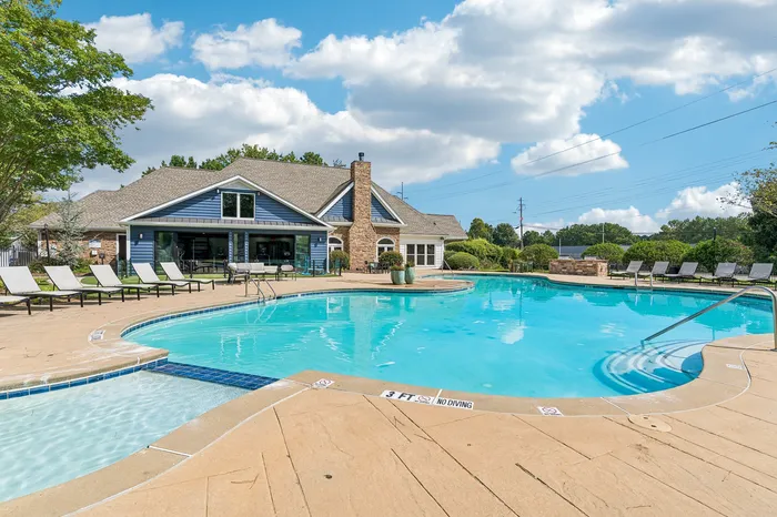 Inviting outdoor swimming pool with elegant lounge chairs and a stylish clubhouse surrounded by lush greenery under a bright blue sky.