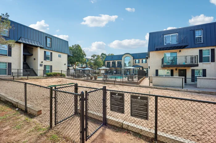 Charming apartment complex with modern architecture, featuring a fenced dog park and inviting pool area under a sunny blue sky.